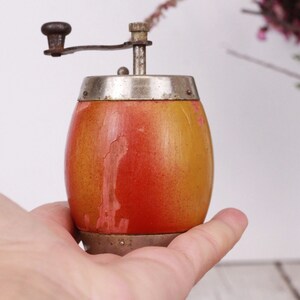 May include: A vintage wooden pepper grinder with a hand crank. The grinder has a barrel-shaped body with a gradient of orange and yellow paint. The top and bottom are silver-colored metal. The crank is dark brown.