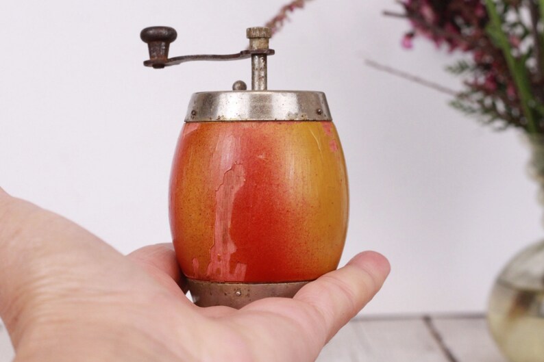May include: A vintage wooden pepper grinder with a hand crank. The grinder has a barrel-shaped body with a gradient of orange and yellow paint. The top and bottom are silver-colored metal. The crank is dark brown.