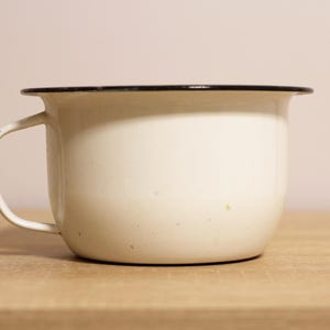 May include: Cream-colored enamel mug with a black rim and handle. The mug sits on a wooden surface. The mug has a rounded shape and a simple design, suitable for beverages.