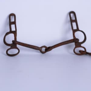Puede incluir: Un bocado de caballo de metal marrón oxidado con una cadena. El bocado tiene una parte superior rectangular con dos rectángulos más pequeños debajo, un anillo circular y una barra curva. La cadena está unida al lado derecho.