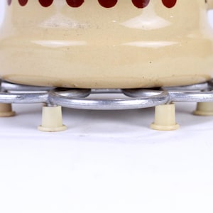 Może przedstawiać: Kremowy emaliowany garnek z wzorem czerwonych i białych kropek stoi na srebrnym metalowym trójnogu z trzema białymi nóżkami.