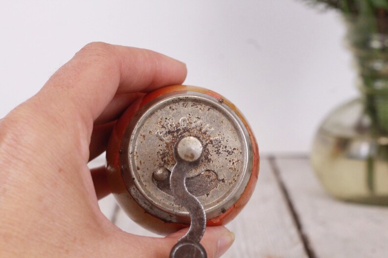 May include: A vintage apple-shaped pencil sharpener with a metal crank and a silver-colored metal base. The sharpener has an orange and yellow body with a slightly worn appearance. The crank is in the down position.