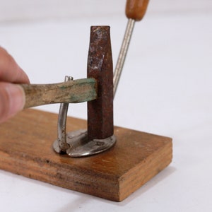 Puede incluir: Una base de madera con un marco de metal y dos martillos de metal oxidados. Un martillo está siendo sostenido por una mano y el otro está descansando sobre la base.