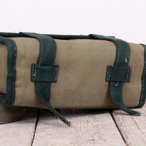 K&ouml;nnte beinhalten: Eine rechteckige olivgr&uuml;ne Canvas-Tasche mit dunkelgr&uuml;nen Zierleisten und Riemen. Die Tasche hat zwei Riemen oben und einen langen Riemen, der sich von der Seite erstreckt. Die Tasche steht auf einer Holzoberfl&auml;che.