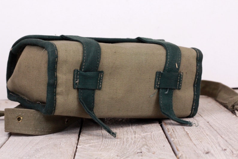 K&ouml;nnte beinhalten: Eine rechteckige olivgr&uuml;ne Canvas-Tasche mit dunkelgr&uuml;nen Zierleisten und Riemen. Die Tasche hat zwei Riemen oben und einen langen Riemen, der sich von der Seite erstreckt. Die Tasche steht auf einer Holzoberfl&auml;che.