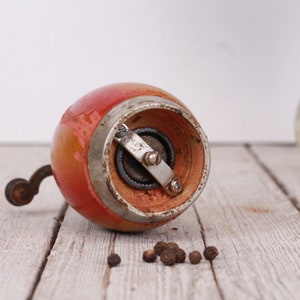 May include: A vintage, hand-cranked pepper mill with a red and orange painted wooden body and a metal grinding mechanism. The mill sits on a weathered wooden surface, with a few peppercorns scattered nearby. A classic kitchen tool.