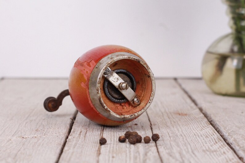 May include: A vintage, hand-cranked pepper mill with a red and orange painted wooden body and a metal grinding mechanism. The mill sits on a weathered wooden surface, with a few peppercorns scattered nearby. A classic kitchen tool.
