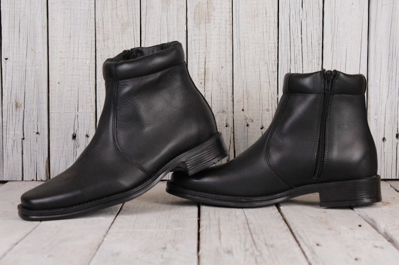 Peut inclure: Une paire de bottines en cuir noir avec un bout carr&eacute; et une fermeture &eacute;clair. Les bottines ont un petit talon et un col rembourr&eacute;. Les bottines sont pr&eacute;sent&eacute;es sur une surface en bois blanc.
