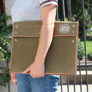 Peut inclure: Une serviette en toile vert olive avec une bordure marron et une fermeture à cordon rouge. Le portfolio porte le numéro "160" dans un cercle blanc sur le devant. Une personne porte le portfolio, vêtue d'un t-shirt gris et d'un jean bleu.