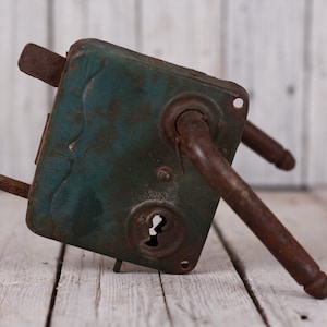 May include: An antique door lock with a teal-colored metal plate and rusty brown handles. The lock has a keyhole and is mounted on a weathered wooden surface, showcasing its vintage aesthetic.