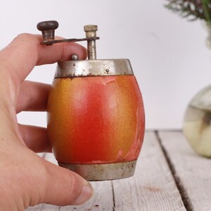 May include: A vintage pepper grinder with a barrel-shaped body. The grinder is painted with a gradient of orange and yellow, and has a silver-colored metal top and bottom. The grinding mechanism is also silver-colored and has a small wooden knob.