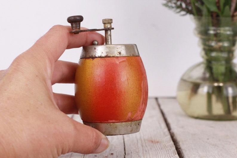 May include: A vintage pepper grinder with a barrel-shaped body. The grinder is painted with a gradient of orange and yellow, and has a silver-colored metal top and bottom. The grinding mechanism is also silver-colored and has a small wooden knob.