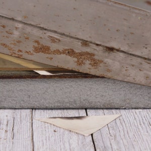 May include: A weathered, gray wooden structure with visible rust and a triangular piece of paper. The wood has a distressed appearance, suggesting age or exposure to the elements. The paper is a light color with a dark stain.