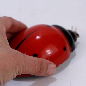 Pode incluir: Um brinquedo de joaninha vermelho e preto com acabamento brilhante. O brinquedo é feito de plástico e tem um mecanismo de corda.
