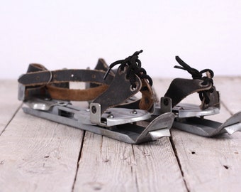May include: A pair of vintage ice skate attachments with black leather straps and buckles. The metal blades are silver and have a worn look.