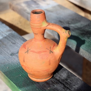 Puede incluir: Una jarra de barro cocido con asa y pico. La jarra está decorada con un diseño rojo y naranja y tiene un patrón de libélula.