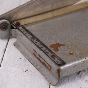 May include: A vintage silver paper cutter with a ruler marked in inches. The cutter has a metal blade and a gray base with some rust spots. The paper cutter is on a white wooden surface.