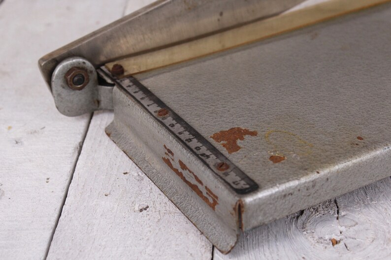 May include: A vintage silver paper cutter with a ruler marked in inches. The cutter has a metal blade and a gray base with some rust spots. The paper cutter is on a white wooden surface.