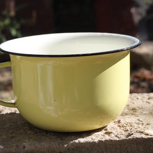 May include: A yellow enamelware cup with a black rim and a handle. The cup is sitting on a rough, textured surface.