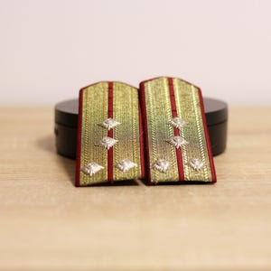 以下が含まれることがあります： 軍の肩章のペア。肩章は、赤いストライプとボーダーが付いた金色の背景です。銀色の星型のエンブレムが肩章に配置されています。肩章は木製の表面に表示されています。