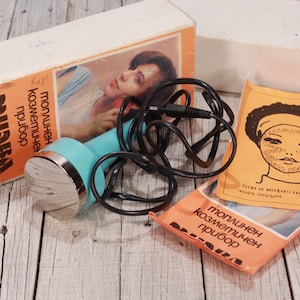 May include: Vintage cosmetic device with a teal handle and silver head, accompanied by a box and pamphlets. The box features an image of a woman and Cyrillic text. A black cord is connected to the device.