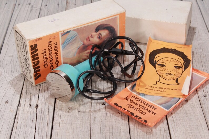 May include: Vintage cosmetic device with a teal handle and silver head, accompanied by a box and pamphlets. The box features an image of a woman and Cyrillic text. A black cord is connected to the device.