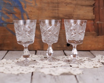 Puede incluir: Tres copas de vino de cristal transparente con intrincados patrones tallados. Las copas tienen una base con tallo y están sobre un tapete de encaje blanco. El fondo es una superficie de madera rústica.