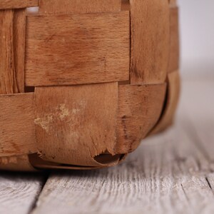 Puede incluir: Primer plano de una cesta tejida con tiras de madera marr&oacute;n claro. La construcci&oacute;n de la cesta presenta un patr&oacute;n entrelazado, con textura visible y ligeras imperfecciones. El borde inferior de la cesta es visible, apoyado sobre una superficie de madera.