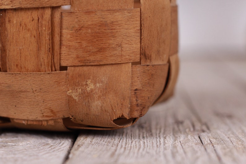 Puede incluir: Primer plano de una cesta tejida con tiras de madera marr&oacute;n claro. La construcci&oacute;n de la cesta presenta un patr&oacute;n entrelazado, con textura visible y ligeras imperfecciones. El borde inferior de la cesta es visible, apoyado sobre una superficie de madera.
