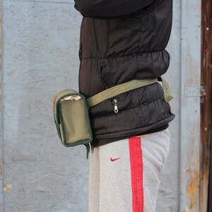 K&ouml;nnte beinhalten: Eine olivgr&uuml;ne Bauchtasche mit passendem G&uuml;rtel wird &uuml;ber einer schwarzen Steppweste getragen. Die Bauchtasche hat eine rechteckige Tasche mit Rei&szlig;verschluss. Die Person tr&auml;gt eine graue Jogginghose mit rotem Streifen.