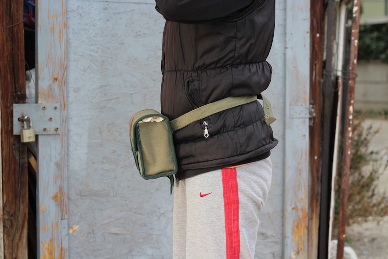 K&ouml;nnte beinhalten: Eine olivgr&uuml;ne Bauchtasche mit passendem G&uuml;rtel wird &uuml;ber einer schwarzen Steppweste getragen. Die Bauchtasche hat eine rechteckige Tasche mit Rei&szlig;verschluss. Die Person tr&auml;gt eine graue Jogginghose mit rotem Streifen.