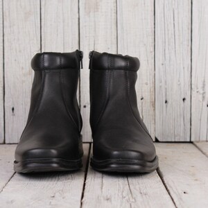 Peut inclure: Une paire de bottines en cuir noir avec fermetures &eacute;clair. Les bottes ont un bout arrondi et un col rembourr&eacute;. Les bottes sont pr&eacute;sent&eacute;es sur une surface en bois blanc.