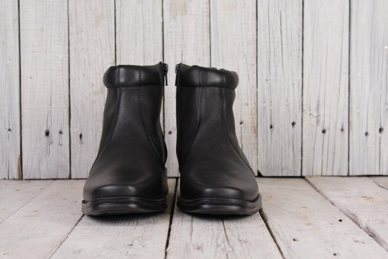 Peut inclure: Une paire de bottines en cuir noir avec fermetures &eacute;clair. Les bottes ont un bout arrondi et un col rembourr&eacute;. Les bottes sont pr&eacute;sent&eacute;es sur une surface en bois blanc.