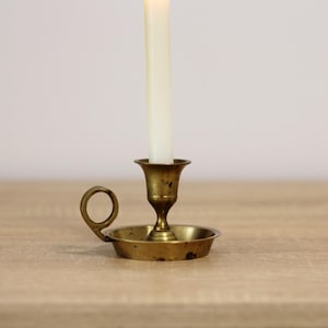 Può includere: Un portacandele vintage in ottone con una candela bianca accesa. Il supporto ha una piccola base rotonda con un manico e una parte superiore a forma di coppa per tenere la candela. La candela è alta e sottile. Il portacandele è su una superficie di legno.