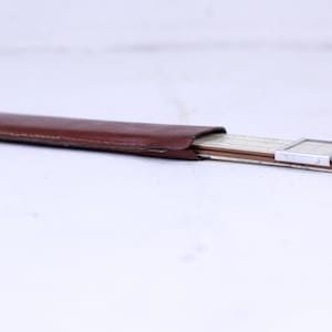 May include: A brown leather case containing a silver slide rule. The slide rule is closed and has a small magnifying glass on the top.