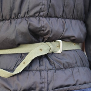 K&ouml;nnte beinhalten: Nahaufnahme einer schwarzen Steppjacke mit einem hellgr&uuml;nen Canvas-G&uuml;rtel. Der G&uuml;rtel hat eine silberfarbene Schnalle und mehrere &Ouml;sen. Die Jacke wird um die Taille getragen.