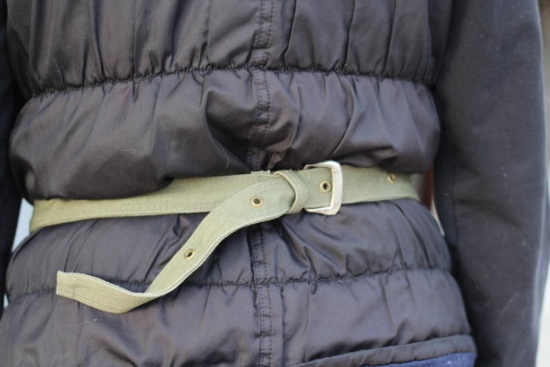 K&ouml;nnte beinhalten: Nahaufnahme einer schwarzen Steppjacke mit einem hellgr&uuml;nen Canvas-G&uuml;rtel. Der G&uuml;rtel hat eine silberfarbene Schnalle und mehrere &Ouml;sen. Die Jacke wird um die Taille getragen.
