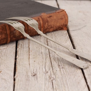 Puede incluir: Dos tenedores de plata con mangos ornamentados se colocan encima de un libro encuadernado en cuero marrón. El libro está sobre una superficie de madera clara.