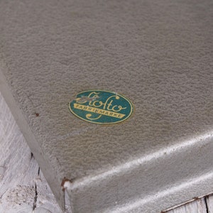 May include: A vintage, textured, gray metal box with a rounded corner. An oval sticker with the text "Otto Fabrikmarke" in green and gold is affixed to the top. The box sits on a white wooden surface.