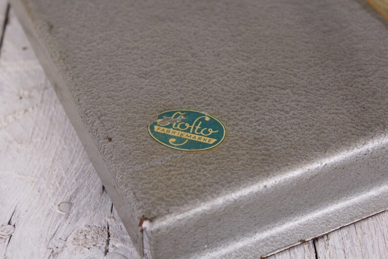 May include: A vintage, textured, gray metal box with a rounded corner. An oval sticker with the text "Otto Fabrikmarke" in green and gold is affixed to the top. The box sits on a white wooden surface.
