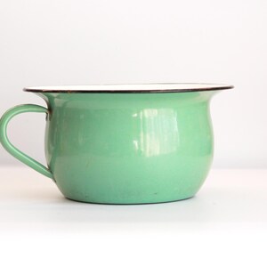 May include: A mint green enamel chamber pot with a smooth, glossy finish and a single curved handle on the side. The vessel features a wide, flared rim with a thin black border, showcasing a rounded, deep basin design for utility.