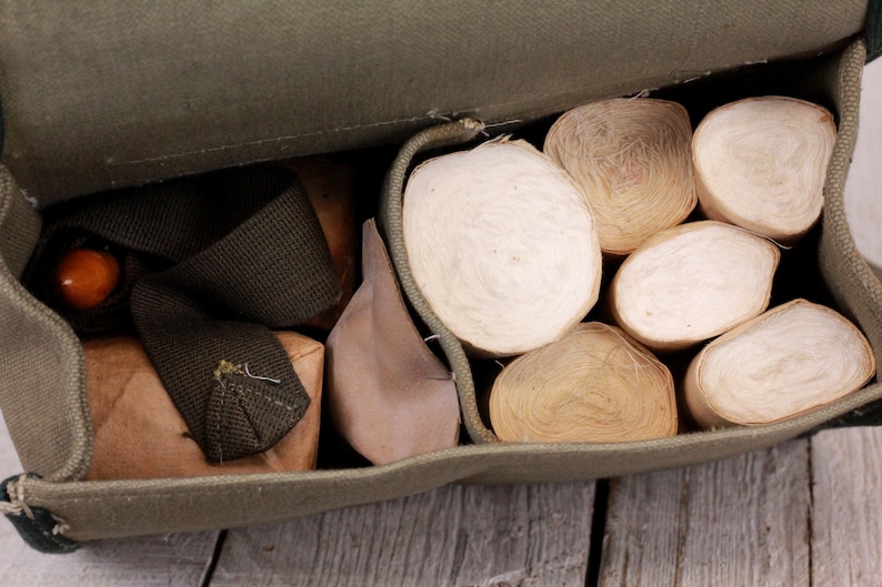 K&ouml;nnte beinhalten: Ge&ouml;ffnete olivgr&uuml;ne Canvas-Tasche, gef&uuml;llt mit mehreren Rollen cremefarbenen Materials. Die Tasche enth&auml;lt auch kleine braune P&auml;ckchen und einen dunkelgr&uuml;nen Riemen. Der Inhalt ist sichtbar.