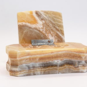 May include: A rectangular box crafted from banded onyx stone in shades of amber, cream, and grey. The box features a hinged lid that stands upright, connected to the base by a silver-toned metal hinge with two visible screws on the top surface.