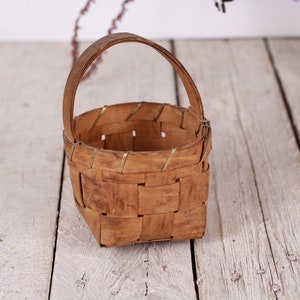 Puede incluir: Una peque&ntilde;a cesta tejida con asa. La cesta est&aacute; hecha de madera marr&oacute;n claro y tiene una base cuadrada. El asa es arqueada y est&aacute; hecha del mismo material. La cesta se encuentra sobre una superficie de madera de color claro.