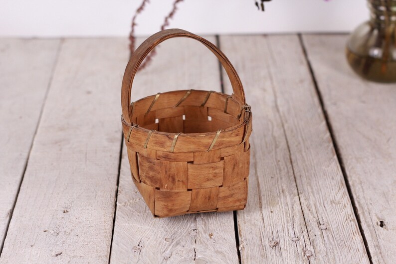 Puede incluir: Una peque&ntilde;a cesta tejida con asa. La cesta est&aacute; hecha de madera marr&oacute;n claro y tiene una base cuadrada. El asa es arqueada y est&aacute; hecha del mismo material. La cesta se encuentra sobre una superficie de madera de color claro.