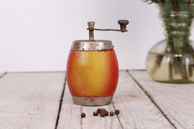 May include: Vintage pepper mill with a wooden body, painted in shades of yellow and red, and accented with silver-toned metal. The mill features a hand crank and sits on a wooden surface with peppercorns scattered nearby.