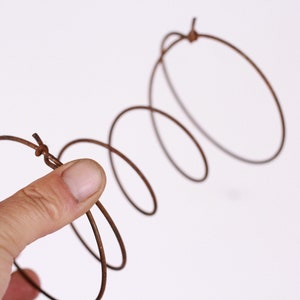 May include: A brown metal spring held by a hand against a white background. The spring is coiled and has a series of circular loops. The metal appears to be aged or weathered, giving it a rustic appearance.