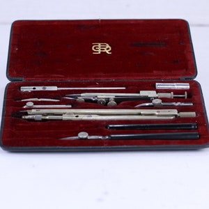 Könnte beinhalten: Ein Satz Zeichenwerkzeuge in einem mit rotem Samt ausgekleideten Etui. Die Werkzeuge umfassen Zirkel, Lineal und Geodreieck. Das Etui trägt eine goldene Inschrift "E.R."