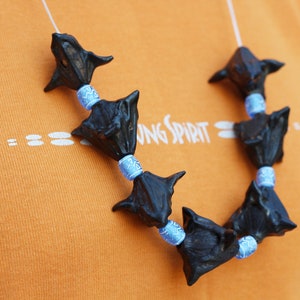 May include: A handmade necklace featuring dark, spiky, black beads interspersed with light blue and white patterned beads. The necklace is displayed against an orange background, with the text "YOUNG SPIRIT" visible.