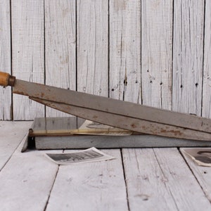 May include: An antique paper cutter with a wooden handle and a metal blade. The cutter is gray and shows signs of wear. Two vintage black and white photographs are on the wooden surface.